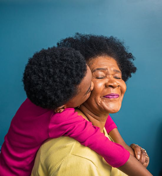 Older woman who is happy while her grandchild wraps their arms around the woman.