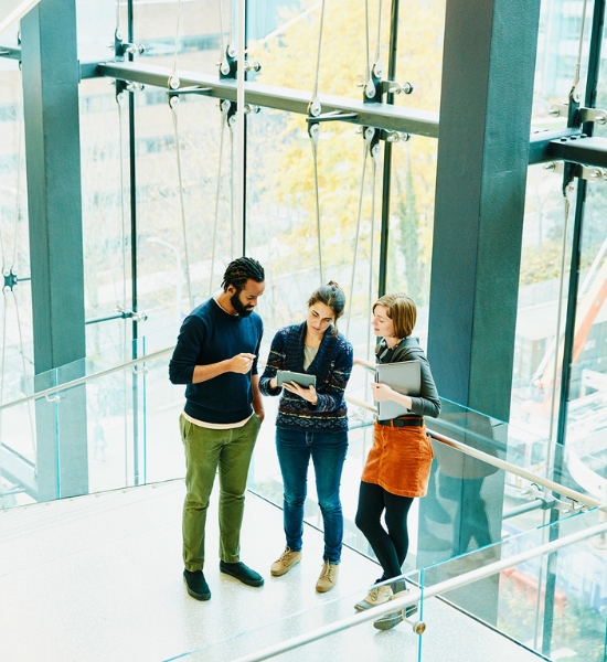 Three colleagues standing in a modern glass office space, reviewing content on a tablet together.
