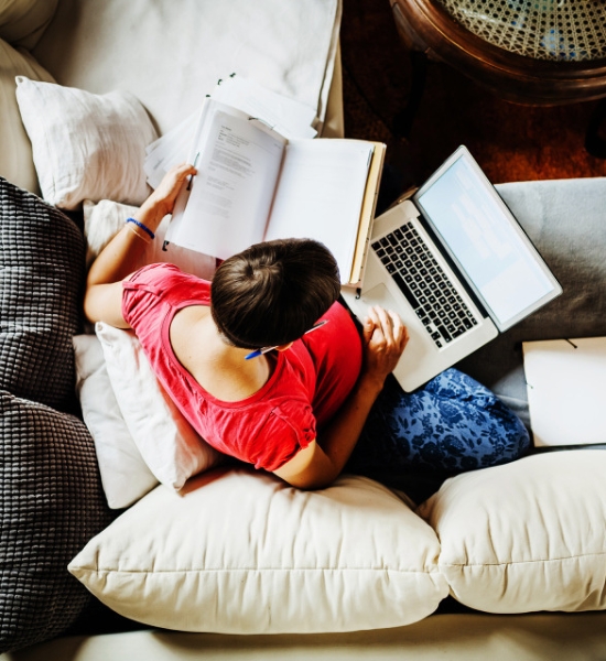 Person relaxing on a couch while reading a book and using a laptop.