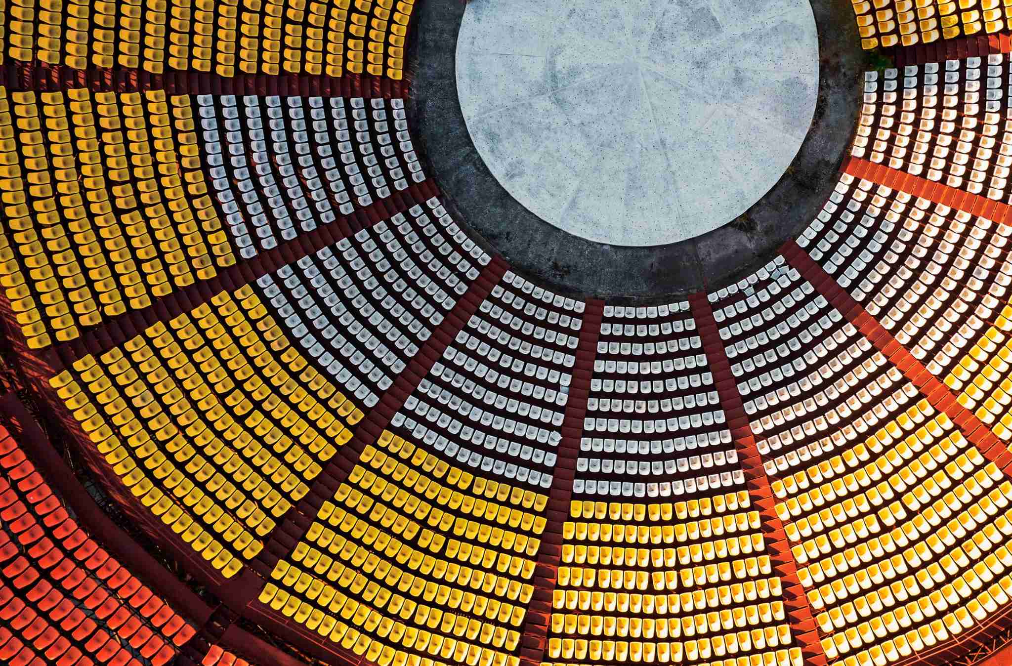 Image shows aerial view of a circular outdoor amphitheater with rows of yellow, white, and red seats arranged in a geometric pattern around a central stage.