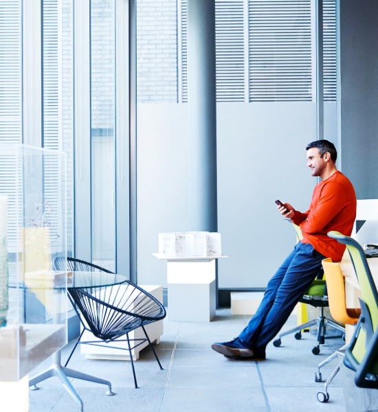 Un uomo vestito in modo informale controlla il suo dispositivo mobile in ufficio, fissando lo sguardo fuori dalla finestra.