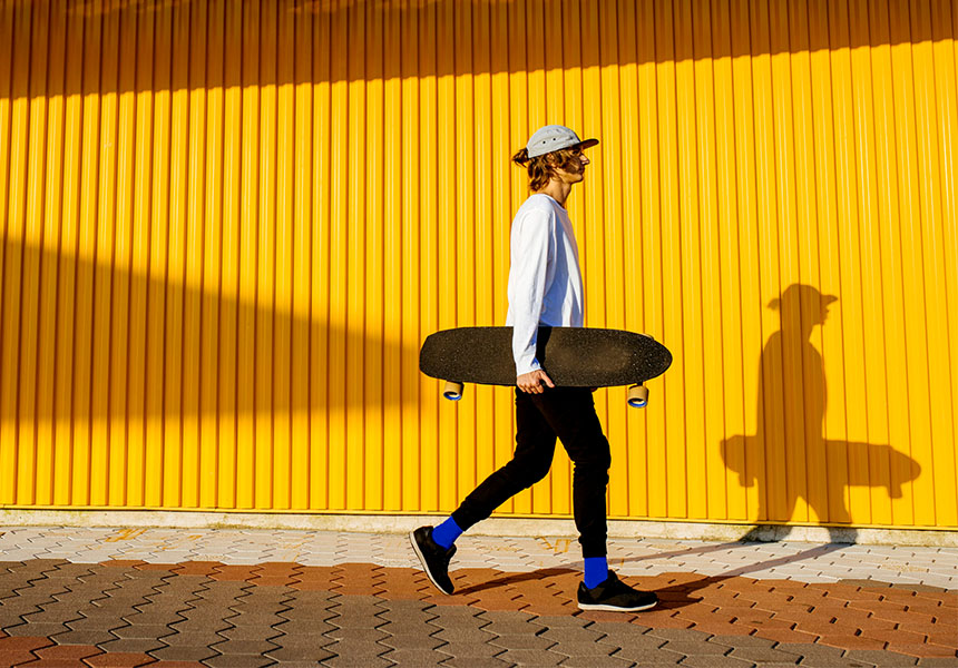 Mladý muž se skateboardem v podpaží kráčí podél žluté zdi.