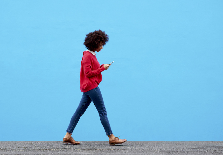 Una mujer camina por la calle, junto a una pared azul claro, mirando su teléfono móvil