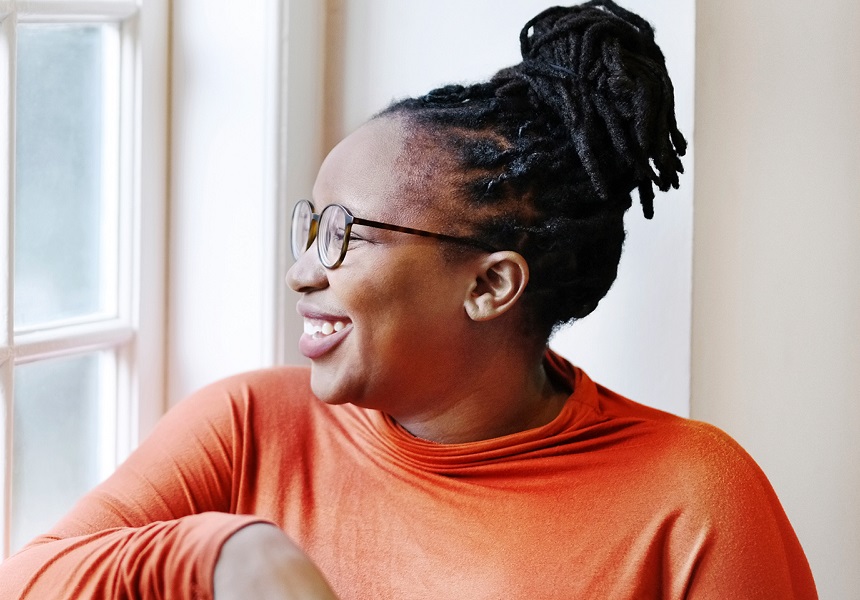 Femme souriante avec des lunettes et des locks, assise près d'une fenêtre en haut orange