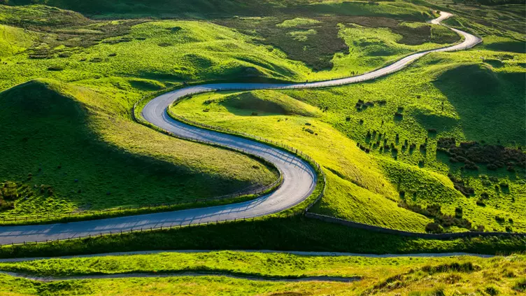 A bendy road which travels down through a bumpy green landscape below Mam Tor and Rushup edge to Edale in the Peak District