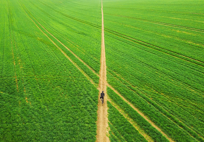 Person walking along a narrow dirt path through a vast green field, viewed from above