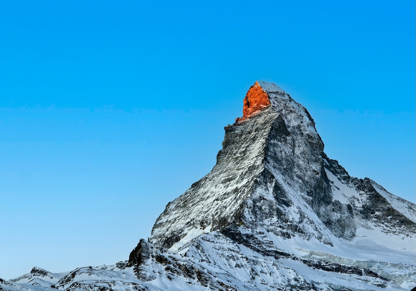 Snow-covered mountain peak with the summit illuminated by orange sunlight against a clear blue sky