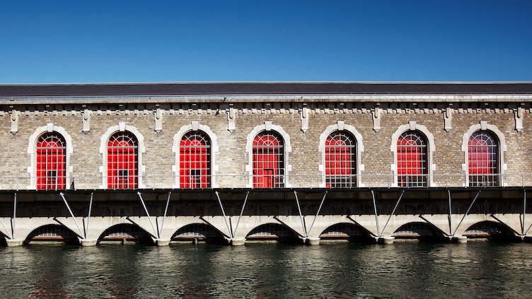Stone building with large arched windows featuring red frames, reflected in the water below under a clear blue sky